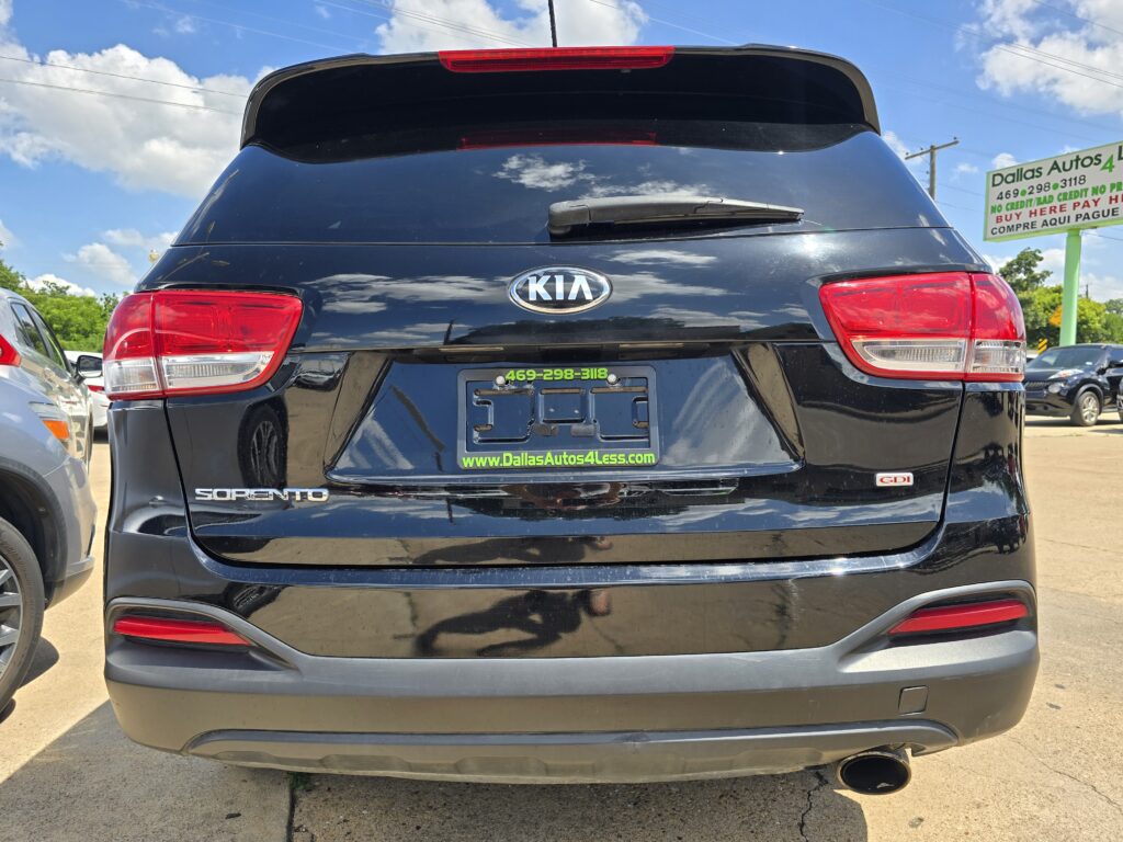 Rear view of a black Kia Sorento SUV at a car dealership, with neon-green dealership branding on the license plate surround and a 'Dallas Autos' sign in the background