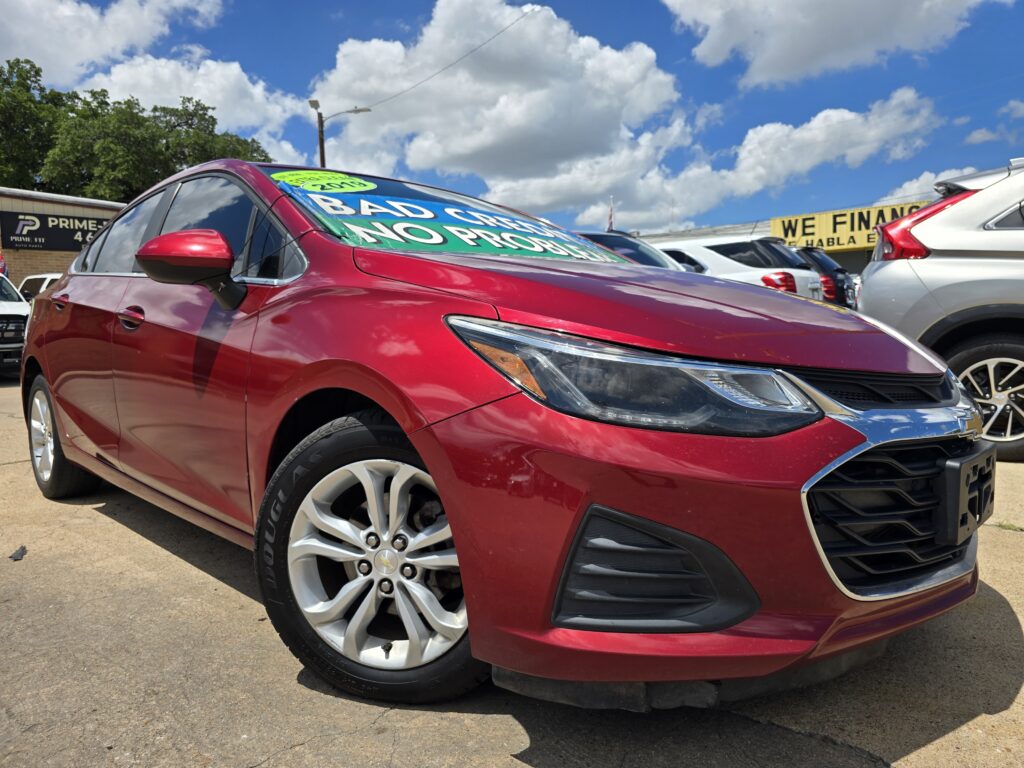 Red Chevrolet sedan parked at a used-car lot with sale signs on the windshield and other cars in the background on a sunny day.