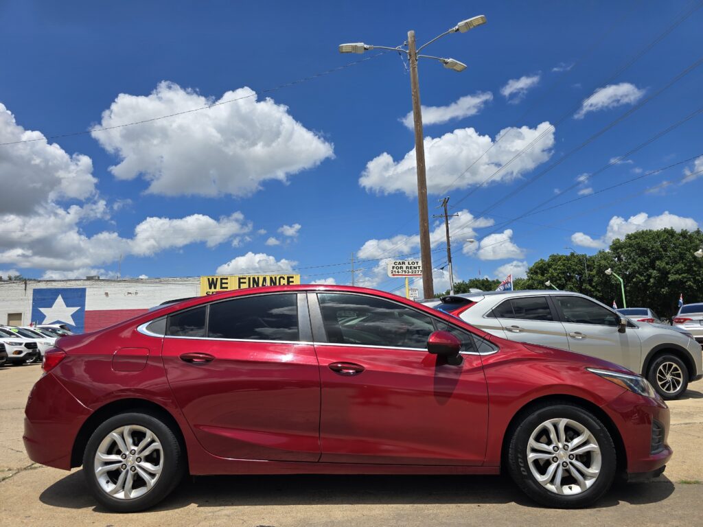 Red sedan parked in a car lot with a large yellow 'WE FINANCE' sign, blue sky and white clouds overhead.