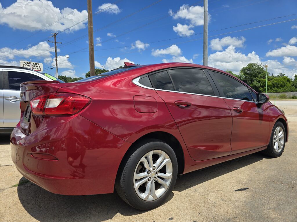 Red sedan parked in a lot with a second car beside it, under a bright blue sky with scattered clouds.