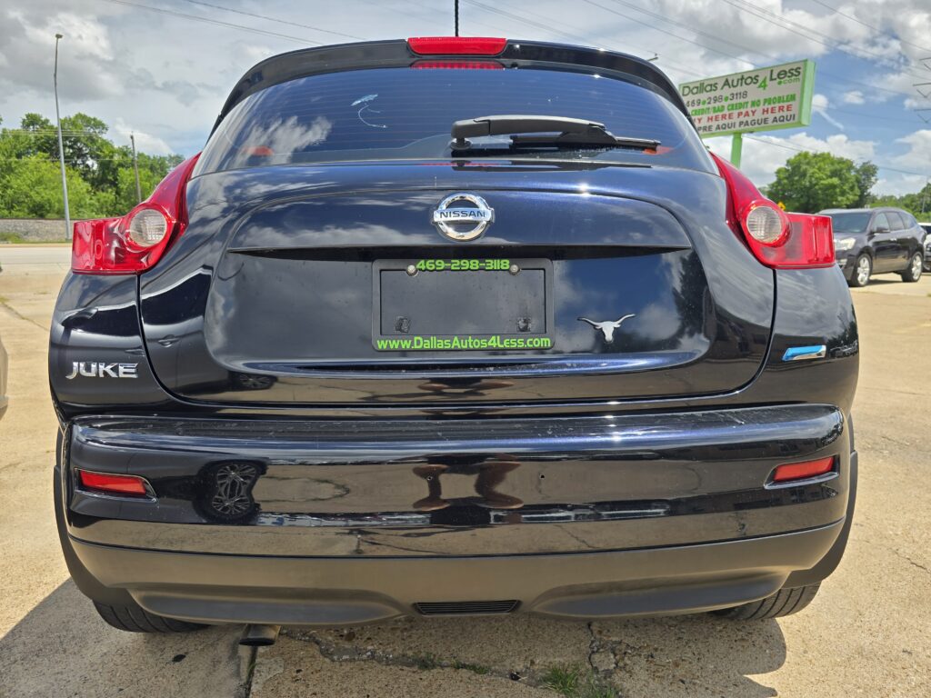 Back view of a black Nissan Juke with a Texas longhorn logo on the bumper, parked at a dealership lot. The plate area shows 'DallasAutos4Less.com' and a phone number.
