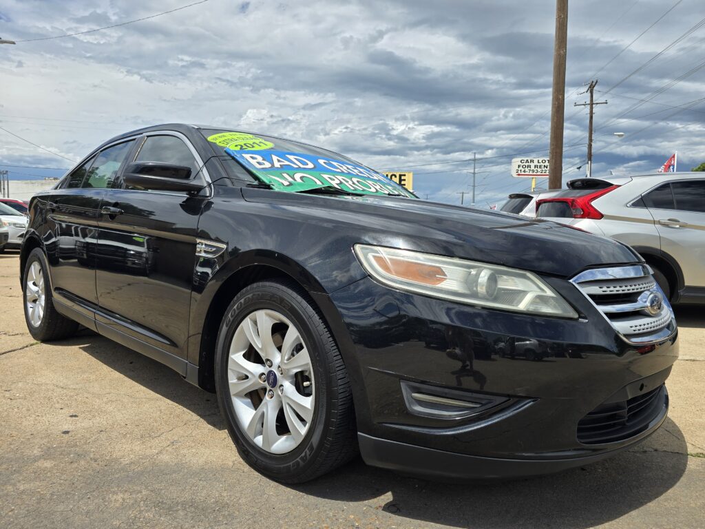 Black Ford sedan in a car lot with a large bright sale sign on the windshield reading “BAD CREDIT” and “2011,” under a cloudy sky.