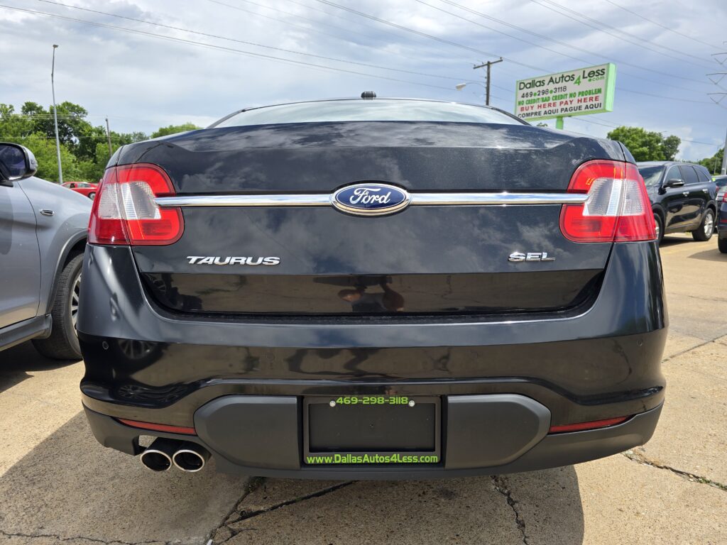 Rear view of a black Ford Taurus SEL in a car dealership lot, showing taillights and dual exhausts with a green Dallas Autos 4 Less sign in the background.