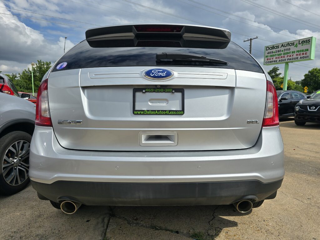Rear view of a silver Ford Edge in a car dealership lot, showing the Ford badge and rear spoiler with a Dallas Autos 4 Less sign in the background