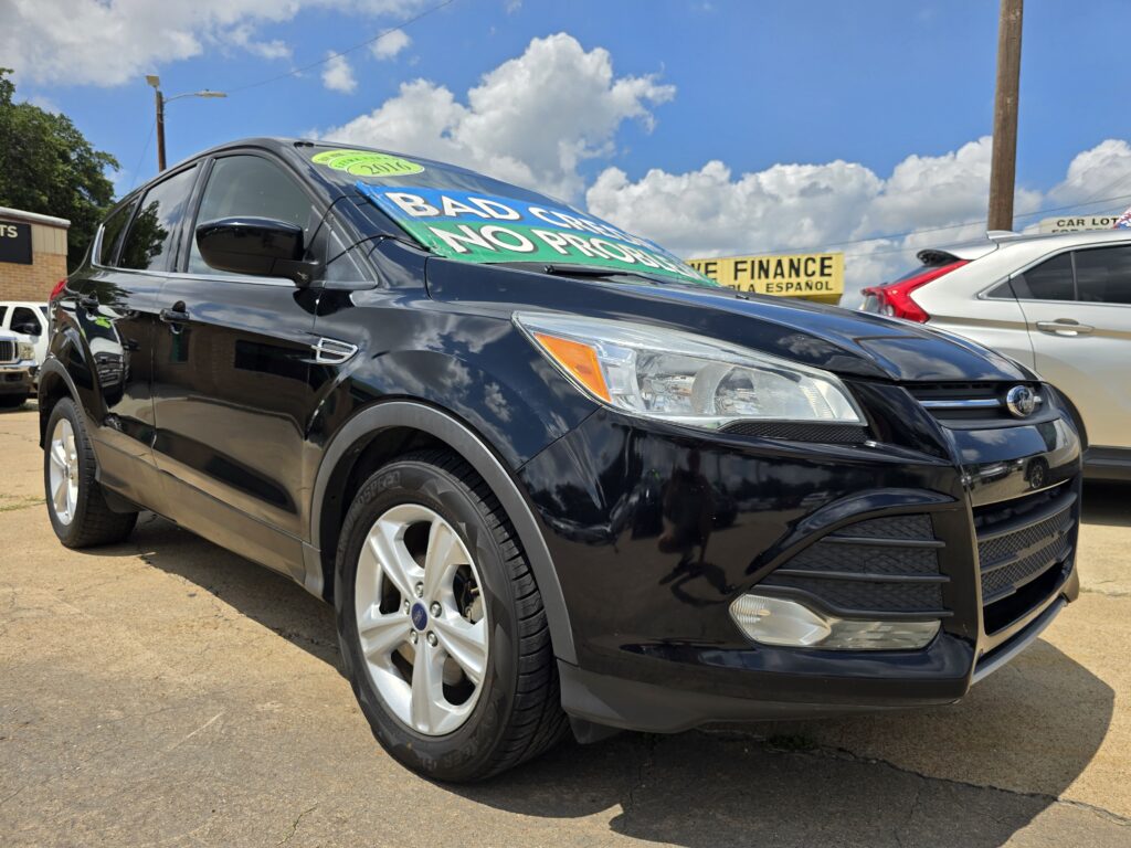 Black SUV parked at a dealer lot with a large green and white sign on the windshield advertising the car for sale. The sky is blue with clouds, and other cars and a finance sign are in the background.