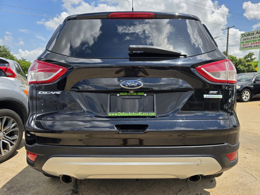 Rear view of a black Ford Escape in a car dealership lot, with a green website URL on the license plate frame and a Dallas Autos sign in the background.