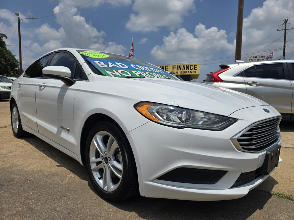 White Ford sedan in a used-car lot with bright green sale signs on the windshield and a blue sky with clouds behind.