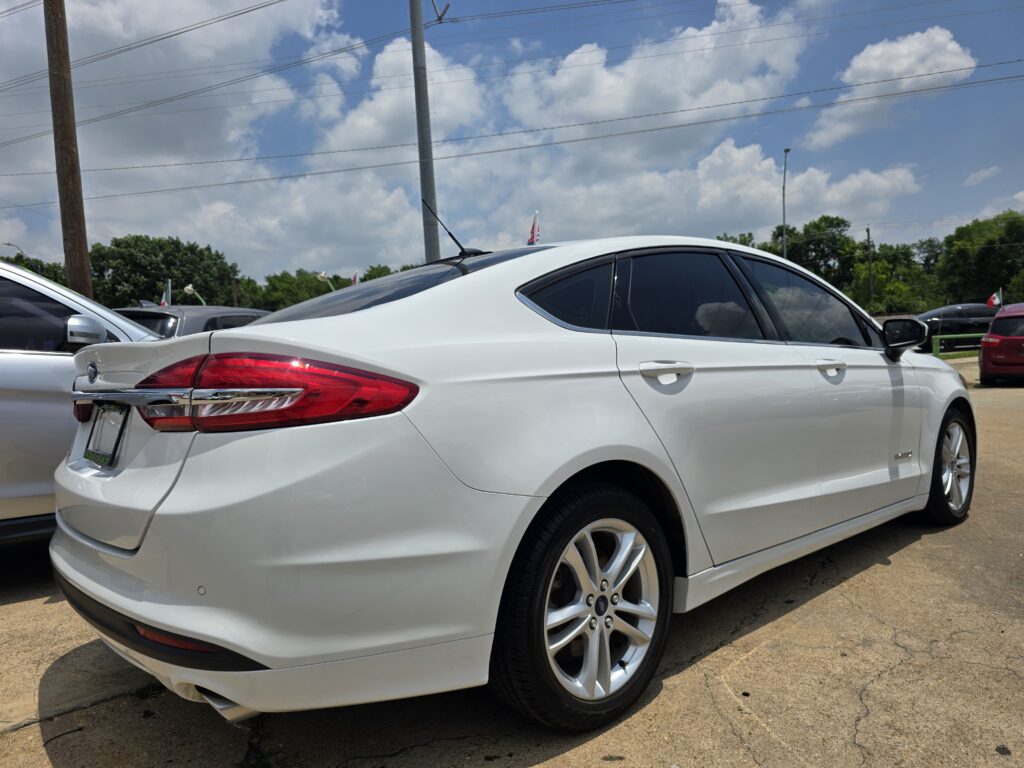 White sedan parked in a lot, showing its rear and side with dark-tinted windows under a blue sky with clouds.