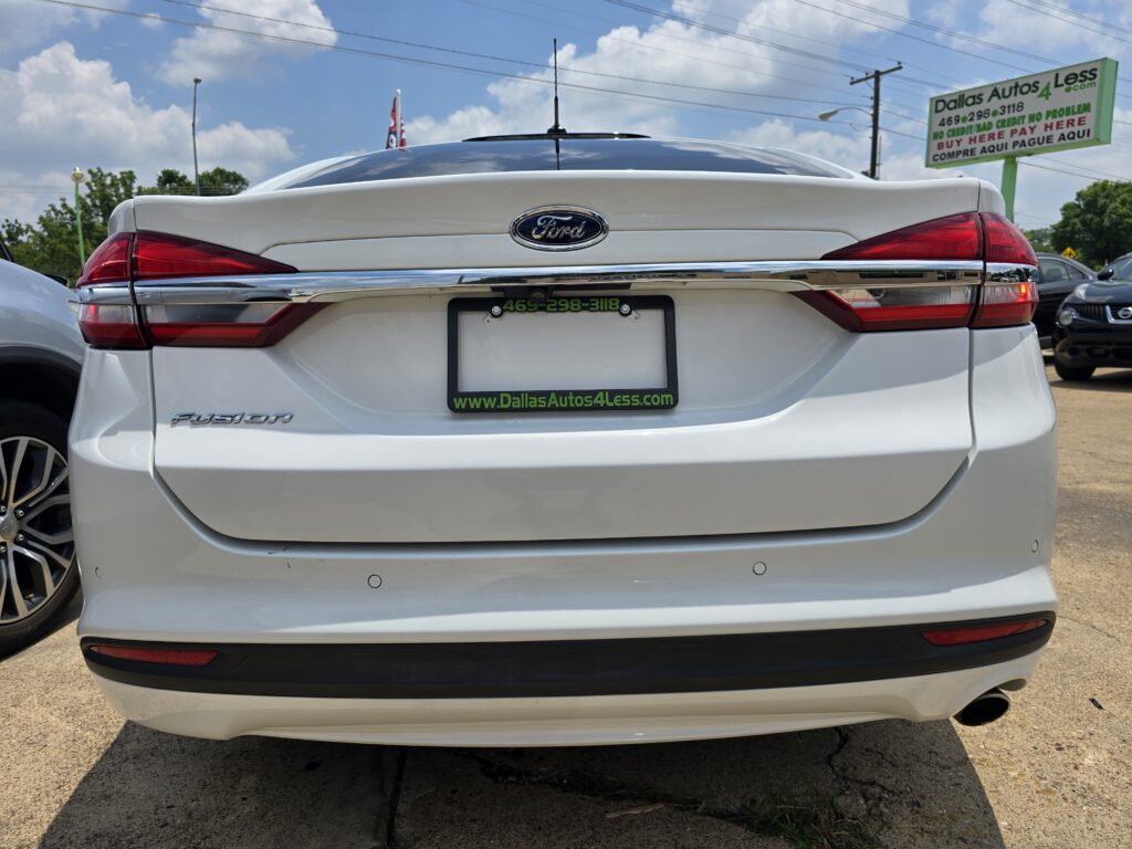 Rear view of a white Ford Fusion with dealership plate frame, at a car lot; big green 'Dallas Autos 4 Less' sign visible in the background