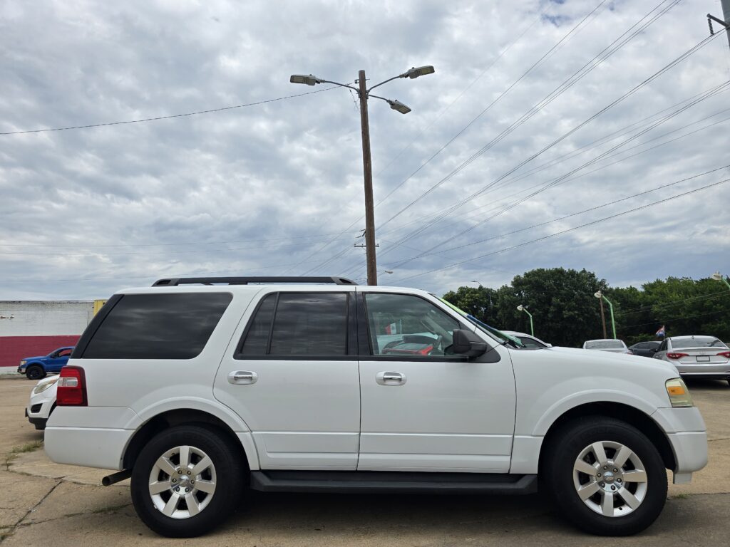 White SUV parked in a lot with a wooden utility pole and multiple streetlights, under a cloudy sky and with other cars and trees in the background.