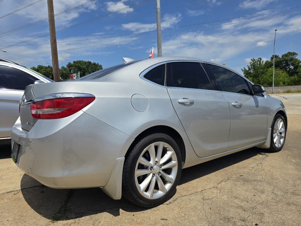 Close-up of a silver sedan parked in a lot, showing rear and passenger side with alloy wheels and red tail light against a blue sky.