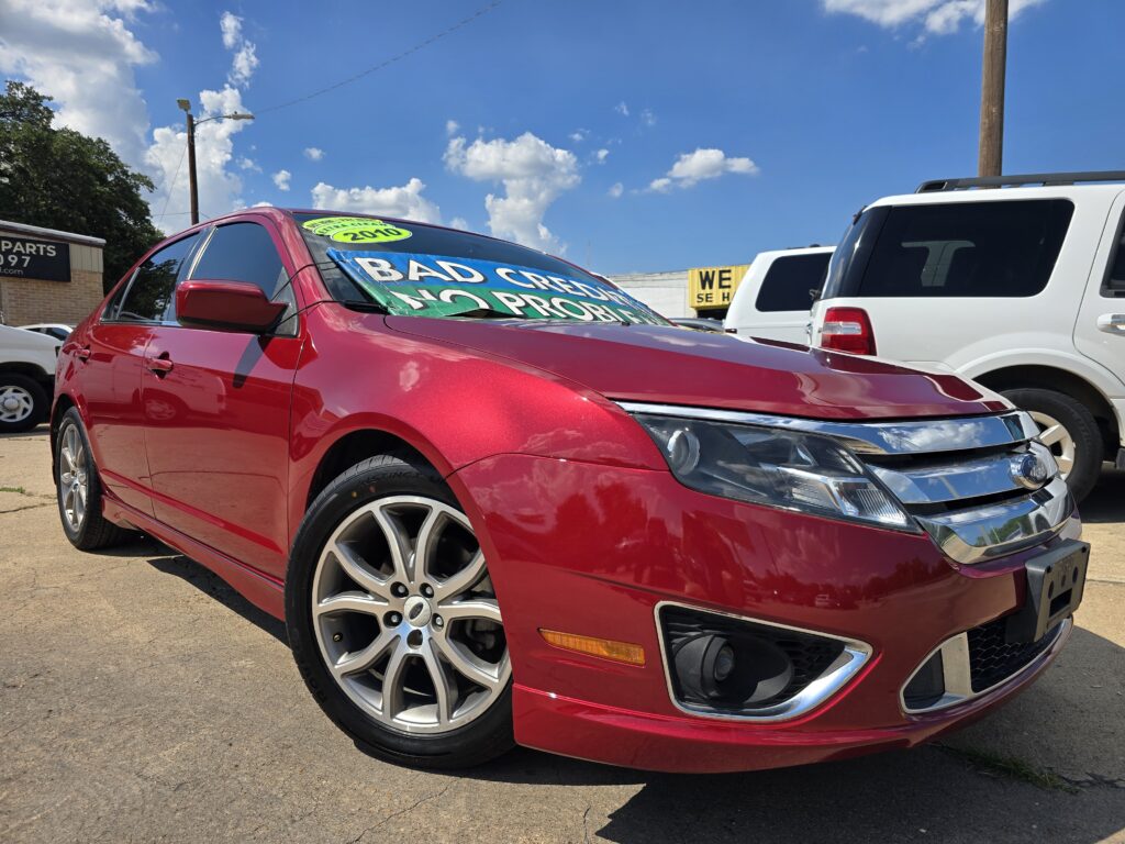 Red sedan in foreground at a used-car lot, with a large sale banner on the windshield and a white SUV in the background under a blue sky.