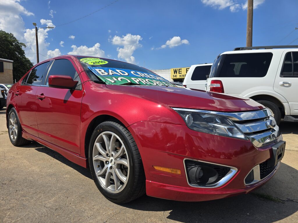 Red sedan in a car dealership lot with large sale banners on the windshield and a white SUV nearby under a blue sky, sunny day.