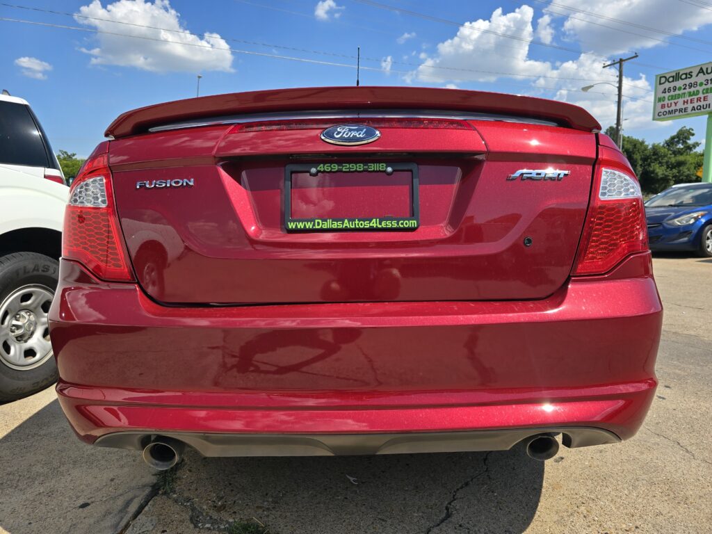 Rear view of a red Ford Fusion with SPORT badge, parked in a lot with a dealership sign in the background