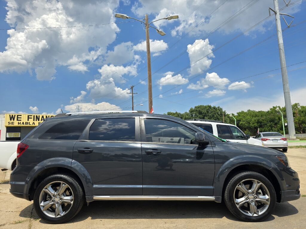 Dark gray SUV parked in a lot with other vehicles, storefront and power poles visible against a blue sky with clouds in the background.