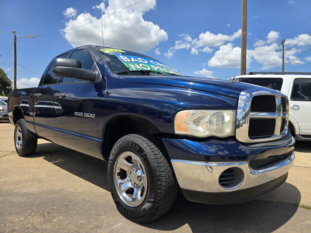 Blue RAM 1500 pickup in a car lot with a green windshield sign offering financing options like 'BAD CREDIT NO PROBLEM'.