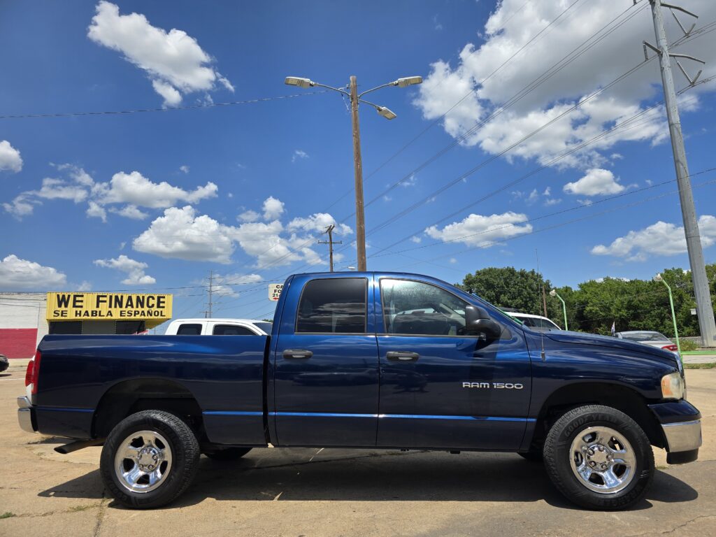 Blue RAM 1500 pickup truck parked in a car lot in front of a yellow WE FINANCE sign on a sunny day.