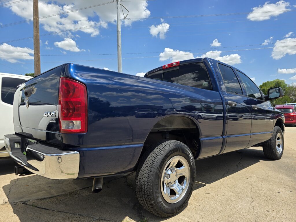 Blue pickup truck rear and side view in a parking lot with clear sky and power lines above, chrome bumper and large wheel visible.