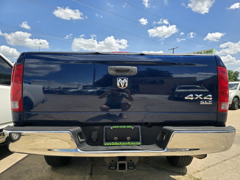 Rear view of a dark blue Ram pickup truck with 4x4 SLT badge, tailgate handle, and hitch, parked at a dealership lot.