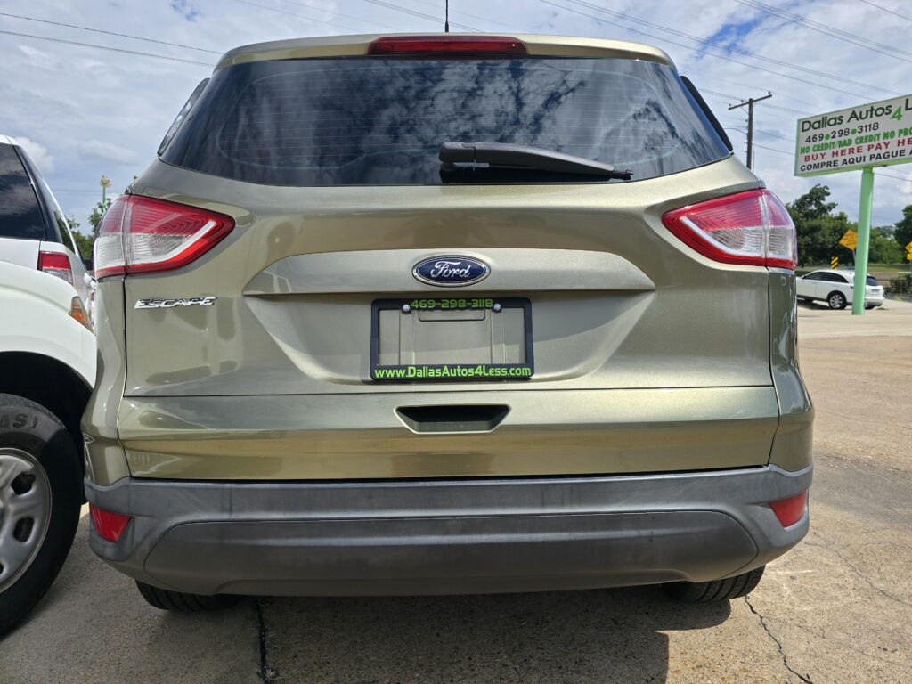 Rear of a gold Ford Escape at a car dealership, with a Dallas Autos4Less sign visible in the background.