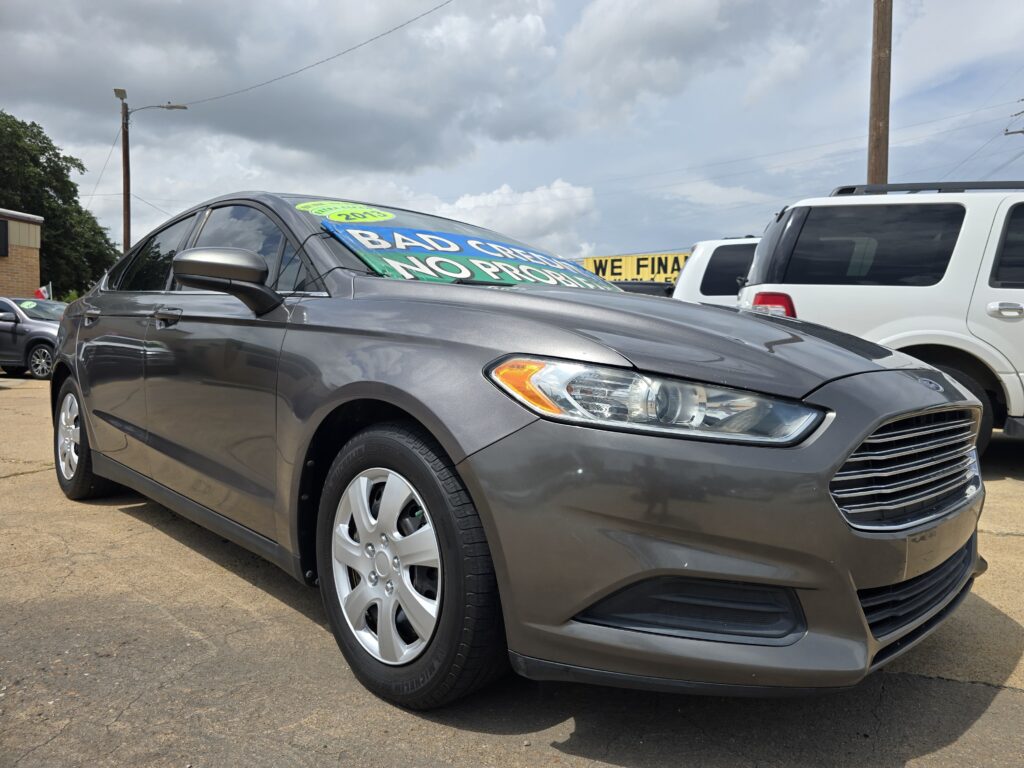 Gray sedan in a car lot with a large green windshield sign saying "BAD CREDIT" and more text, sunny cloudy sky above, other vehicles nearby.