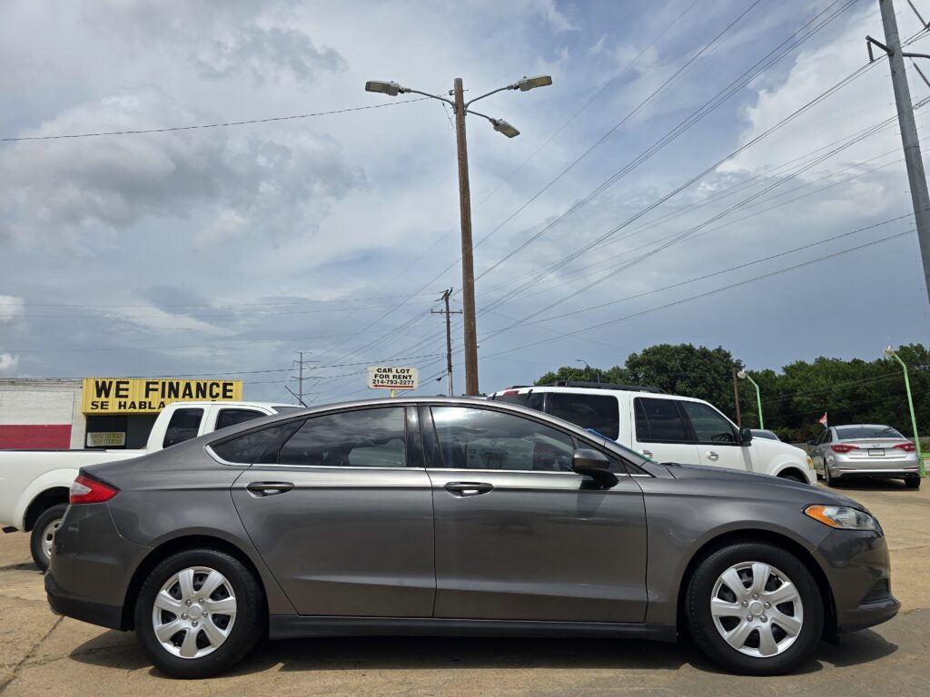 Car dealership lot with several gray cars in the foreground and a yellow 'WE FINANCE' sign on a building in the background.