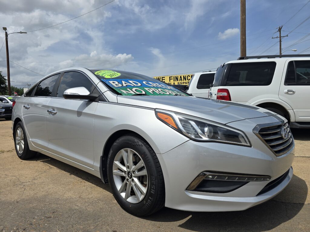 Silver car front and side in a dealership lot with a bright green 2015 sign on the windshield; other vehicles and a yellow sign in the background saying 'WE FINAN...'.