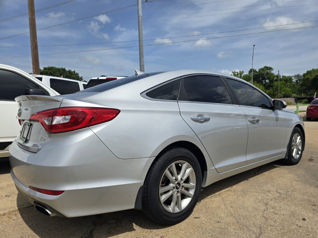 Side/rear view of a silver Hyundai sedan parked in a lot, with other cars and utility poles in the background under a blue sky.