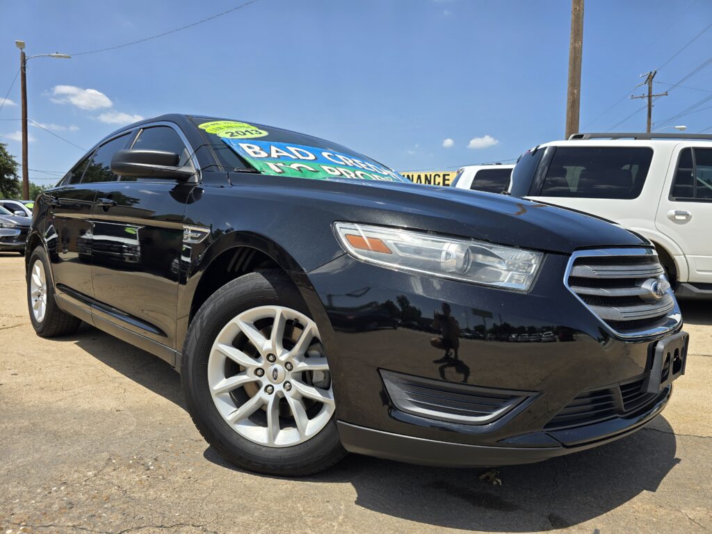 Close-up of a black SUV parked at a dealership, with a bright windshield sign that reads “BAD CREDIT” and other promotional text visible under a blue sky.