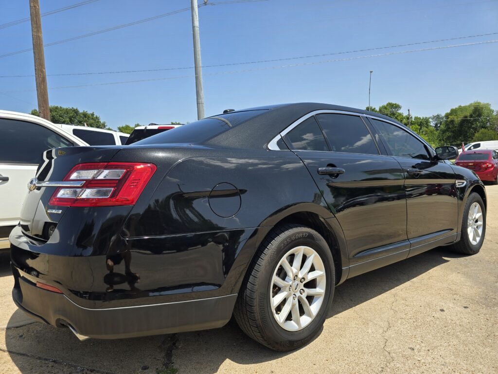 Black sedan parked in a lot, showing rear door, taillight, and chrome trim with other cars and power lines in the background.