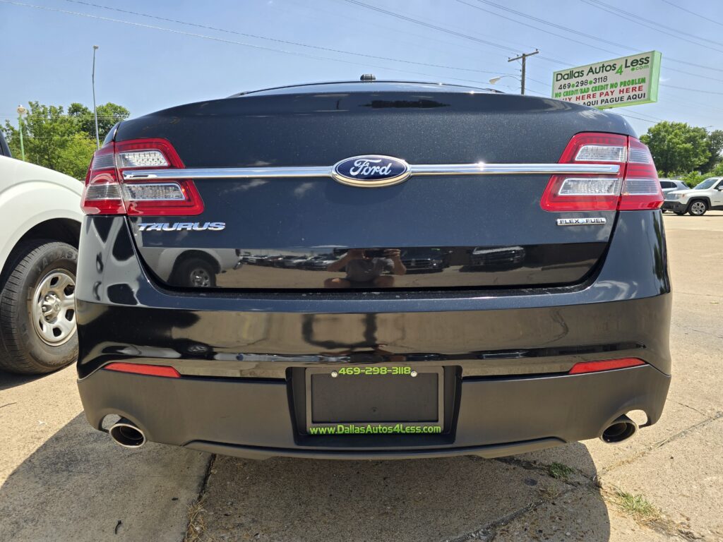 Rear view of a black Ford Taurus showing taillights, Taurus badge, and Flex Fuel emblem in a dealership lot with a Dallas Autos 4 Less sign nearby.