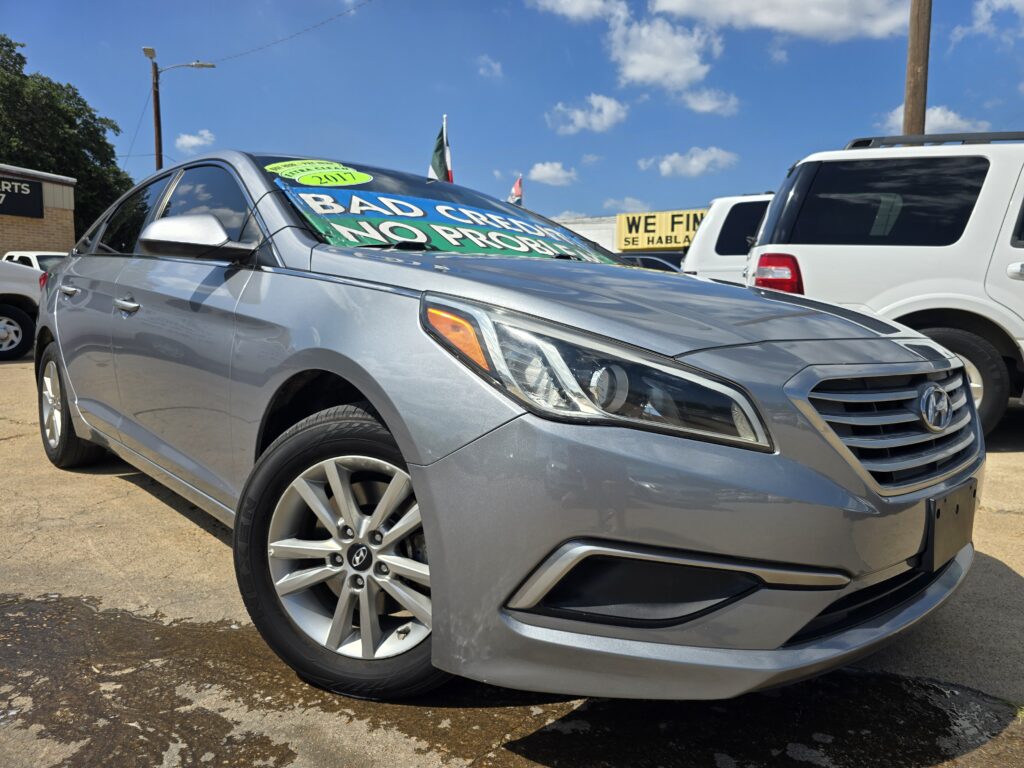 Front corner of a silver Hyundai sedan parked in a lot, with bright green sale signs covering the windshield and a yellow sign in the background under a blue sky.