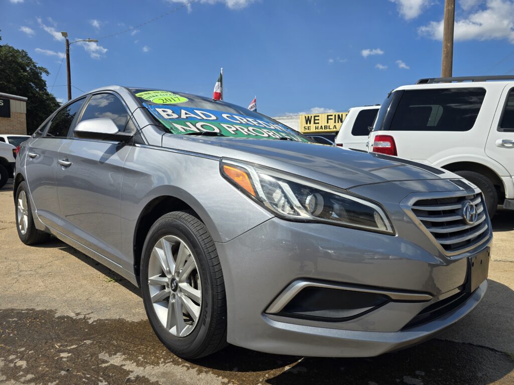 Metallic gray Hyundai sedan with large green 'BAD CAR' signs on the windshield at a sunny used-car lot.