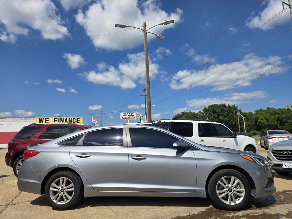 Car dealership/parking lot full of new and used cars under a bright blue sky with power lines and a 'WE FINANCE' sign in the background.