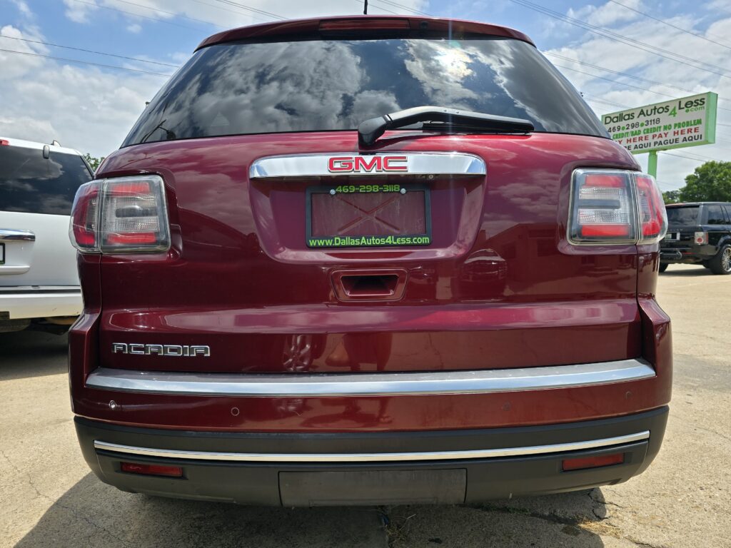Red GMC Acadia rear view with chrome badge and tail lights visible; dealership plate frame and sign in background say Dallas Autos 4 Less .