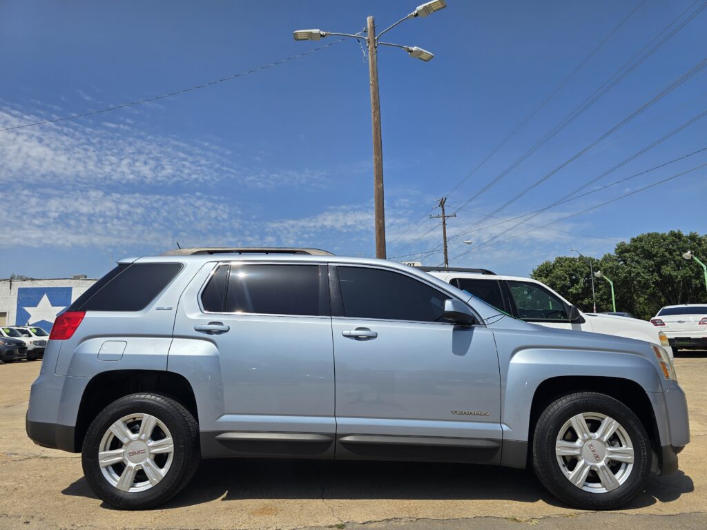 Side view of a silver GMC Terrain SUV in a parking lot, with a wooden utility pole and power lines against a blue sky.