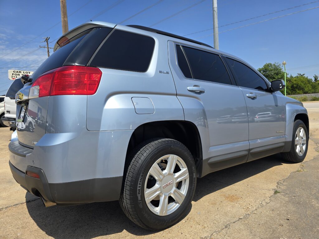 Rear-side view of a light blue GMC SUV with tinted windows and roof rails parked on a sunny street.