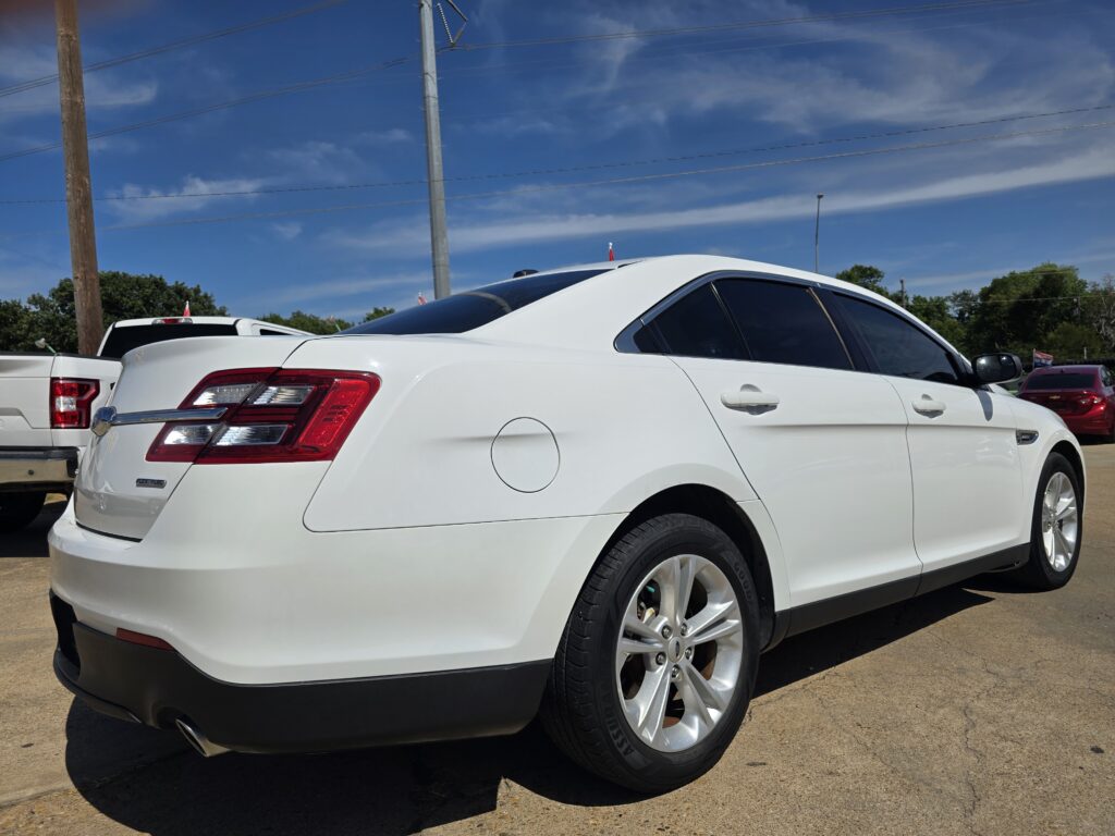 Rear three-quarter view of a white sedan parked in an outdoor lot, with other vehicles and a blue sky overhead.
