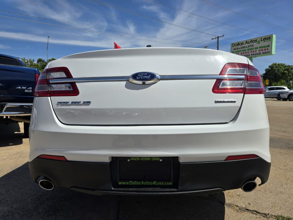 Rear view of a white Ford Taurus in a car dealership lot, with a Dallas Autos 4 Less sign in the background and a blue sky behind.