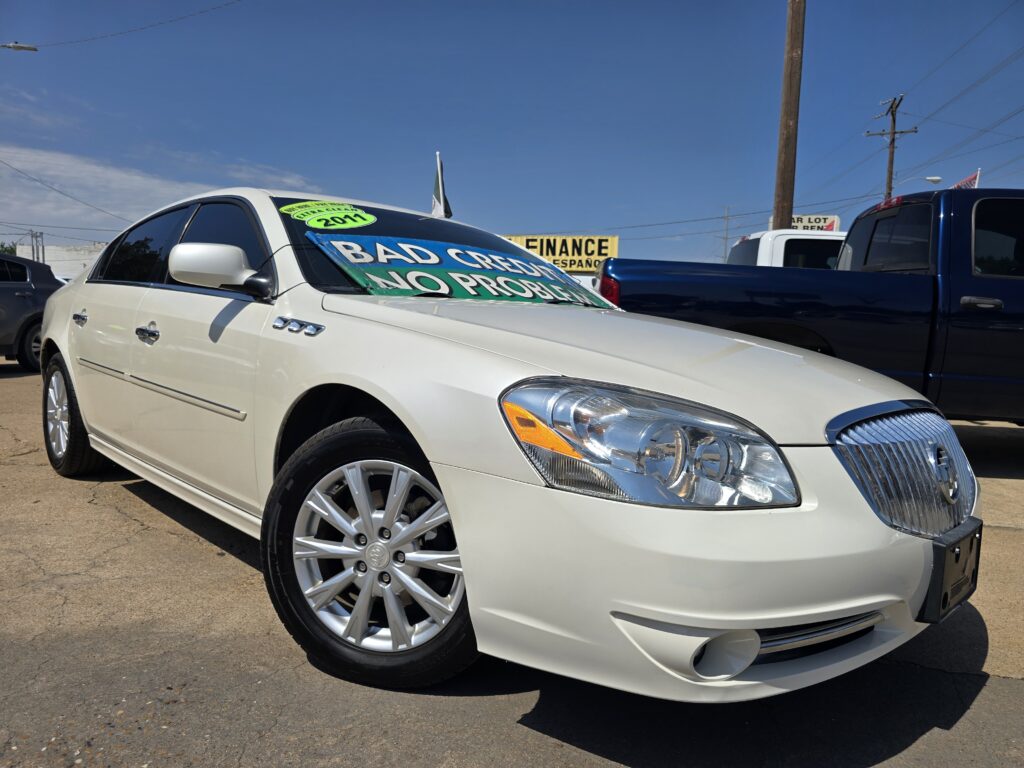 White sedan with large green/blue windshield signs in a used-car lot, reading 'BAD CREDIT NO PRO...' under a clear blue sky.