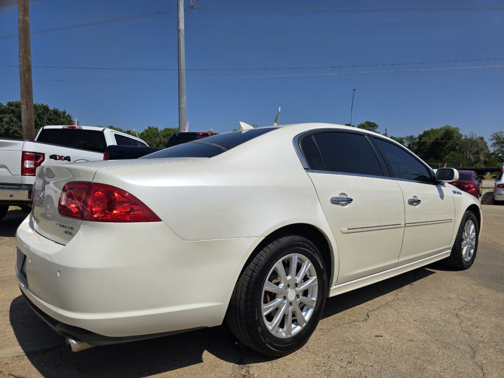 Rear and side view of a white sedan parked in a lot with other vehicles and a blue sky above.
