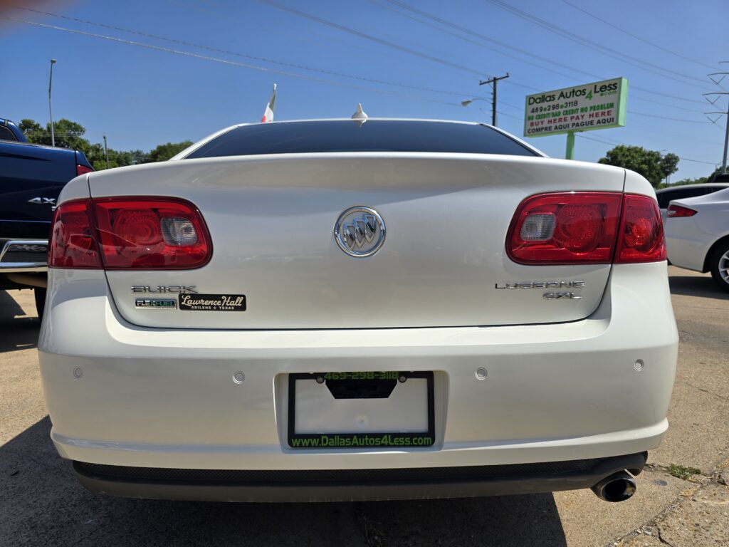 Rear view of a white Buick Lucerne in a dealership lot; red taillights and Buick emblem visible with a Dallas Autos 4 Less sign in the background.