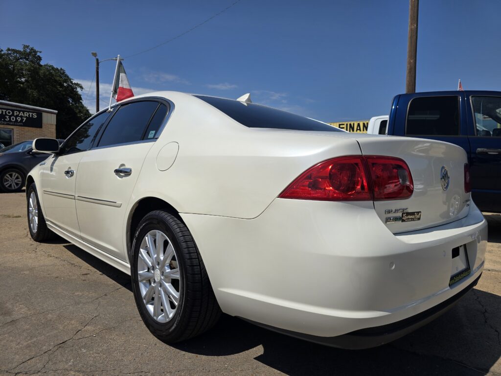 White Buick sedan in a car lot with a US flag mounted on the trunk and blue sky above, dealership sign visible in the background