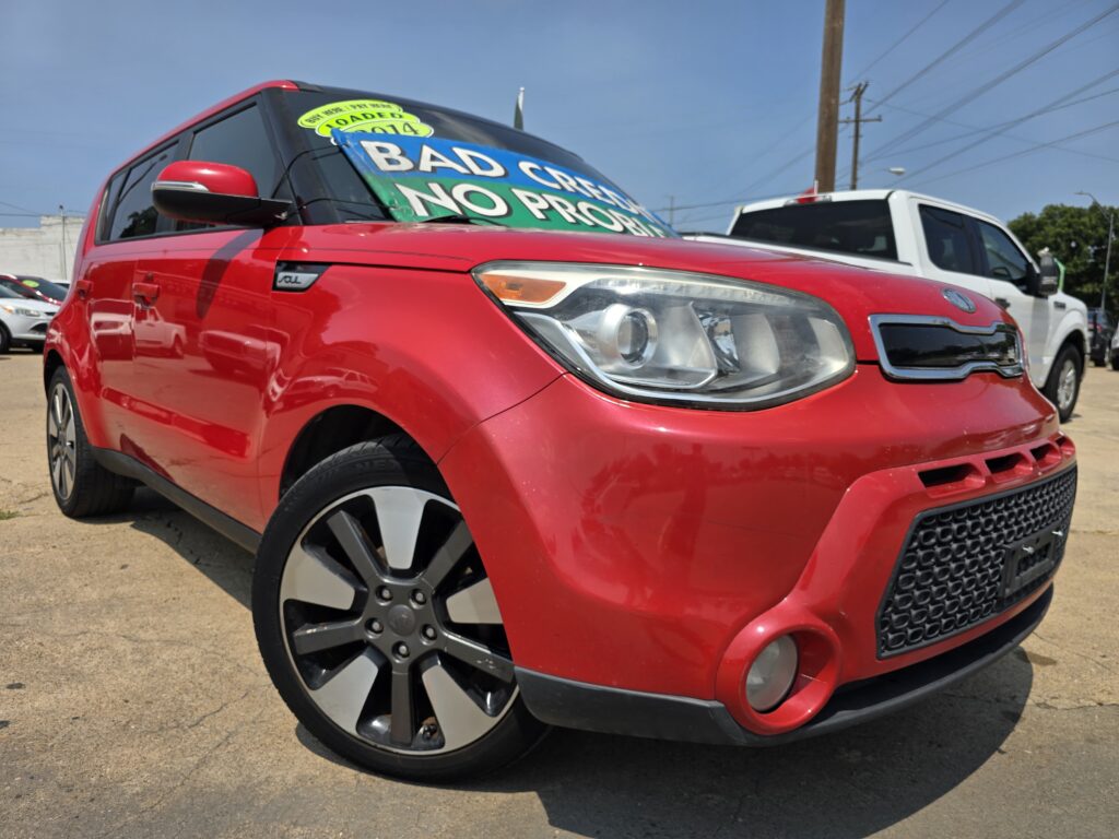 Close-up of a red Kia SUV on a dealership lot with a large windshield banner reading 'BAD CREDIT NO PROBLEM' and a green '2014' sticker nearby.