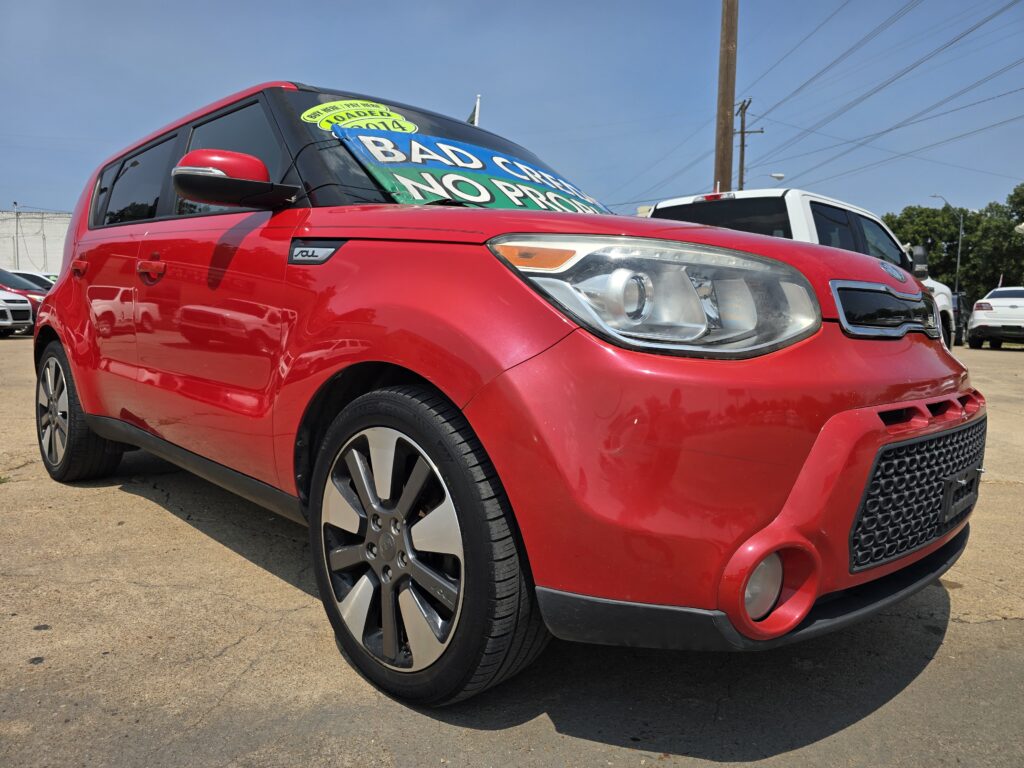 Red SUV at a car dealership with a large sale sign on the windshield reading 'Bad Cars' or similar text, blue sky in the background.