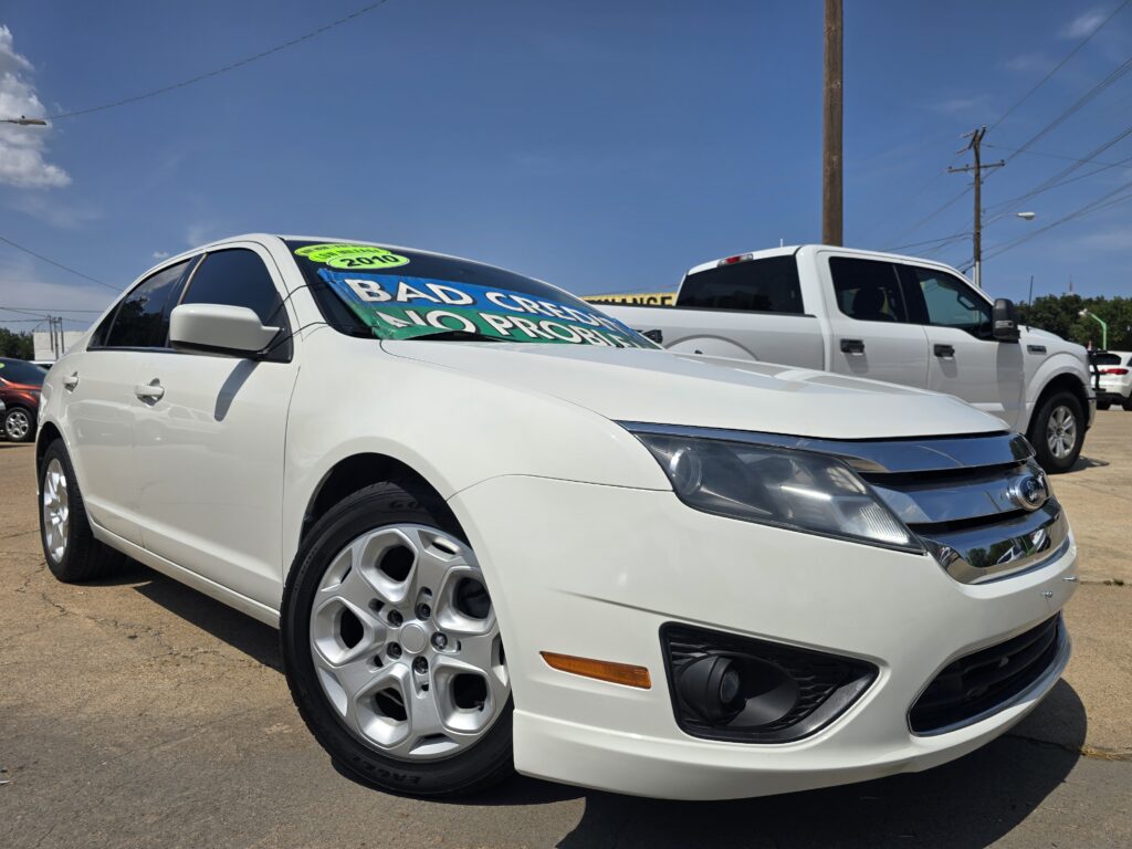 White SUV in foreground with a large green sale banner on the windshield at a sunny car dealership lot. Behind are more white vehicles and power lines under a blue sky.