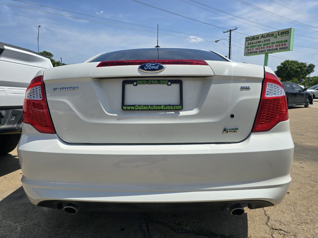 Rear view of a white Ford Fusion SE in a dealership lot, with green Dallas Autos 4 Less license plate frame and red taillights visible.