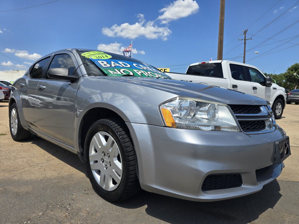 Close-up of a silver sedan with a green windshield sign that reads 'Bad Credit No Problem' at a car lot under a blue sky.