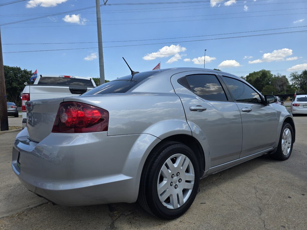 Silver sedan photographed from the rear side in a parking lot, with other vehicles and power lines against a bright blue sky in the background.