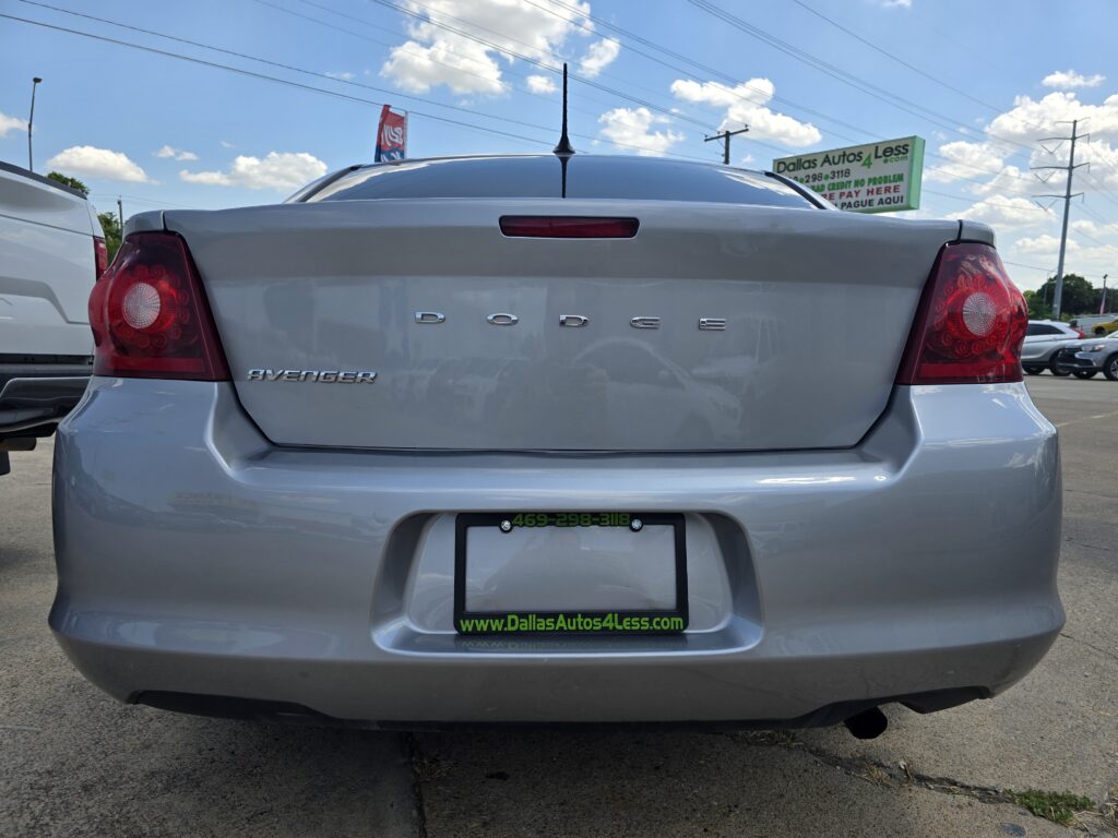 Rear view of a silver Dodge Avenger in a car dealership lot, with a green Dallas Autos 4 Less sign in the background and visible license plate frame reading the site URL.